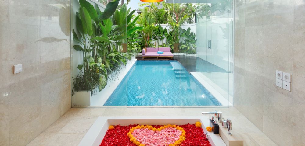 Luxurious bathroom view of a pool through glass. Tub filled with red petals and a heart of pink and yellow. Tropical plants and pink sofa outside. Cozy ambiance.