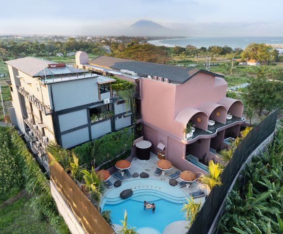 Aerial view of a modern, colorful hotel with a rooftop pool. Nearby are lush green fields and a distant ocean. A relaxed, tropical atmosphere prevails.