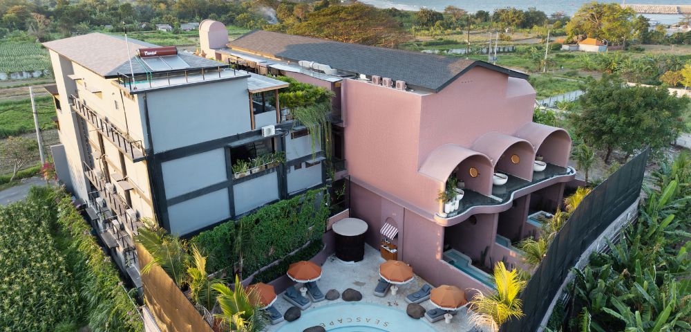 Aerial view of a modern, colorful hotel with a rooftop pool. Nearby are lush green fields and a distant ocean. A relaxed, tropical atmosphere prevails.