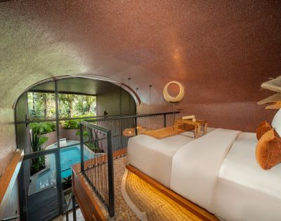 Modern hotel room with a cozy bed, warm lighting, and orange accents. A large window overlooks a lush garden with a serene blue pool, creating a tranquil ambiance.