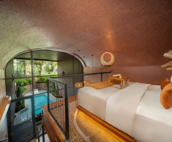 Modern hotel room with a cozy bed, warm lighting, and orange accents. A large window overlooks a lush garden with a serene blue pool, creating a tranquil ambiance.