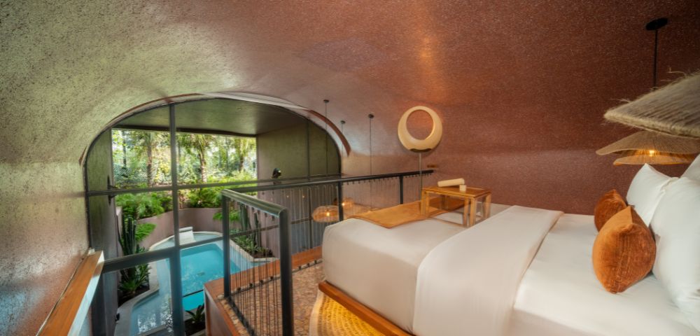 Modern hotel room with a cozy bed, warm lighting, and orange accents. A large window overlooks a lush garden with a serene blue pool, creating a tranquil ambiance.