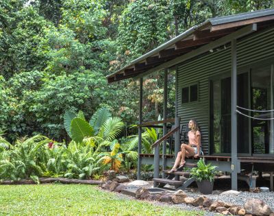 A woman sits on the steps of a modern, corrugated metal cabin surrounded by lush greenery in a tropical setting.
