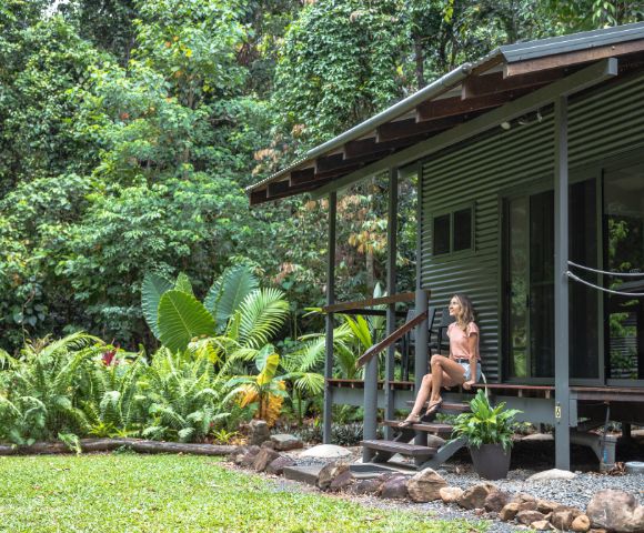 A woman sits on the steps of a modern, corrugated metal cabin surrounded by lush greenery in a tropical setting.