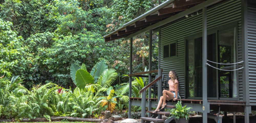 A woman sits on the steps of a modern, corrugated metal cabin surrounded by lush greenery in a tropical setting.