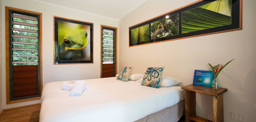 A stylish bedroom featuring a double bed with decorative pillows, framed nature photographs on the wall, wooden furniture, and open windows allowing natural light and greenery to come through.