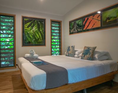 A cozy bedroom featuring a large bed with decorative pillows, two rolled towels, wooden furniture, and green leaf-themed artwork on the walls, with natural light filtering through the slatted windows.