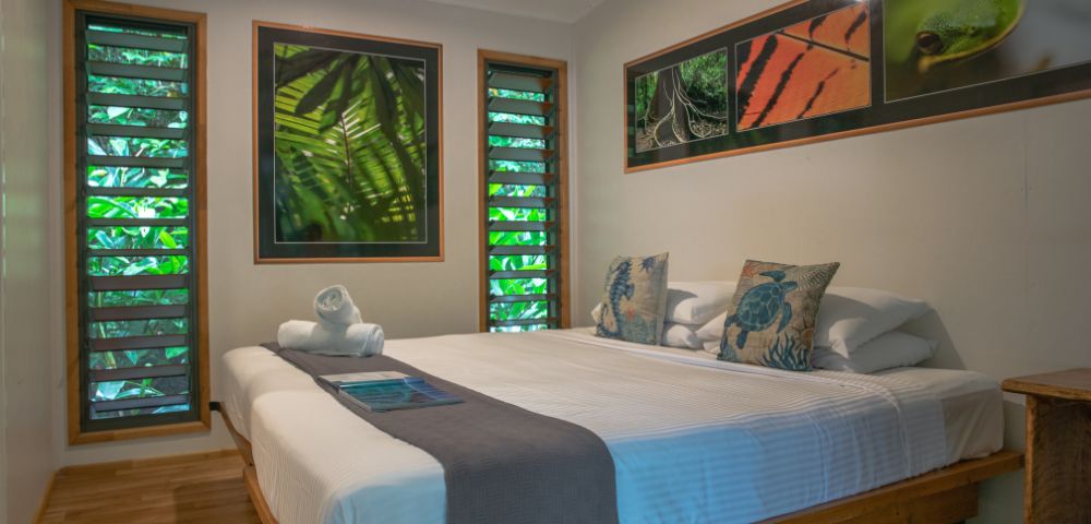 A cozy bedroom featuring a large bed with decorative pillows, two rolled towels, wooden furniture, and green leaf-themed artwork on the walls, with natural light filtering through the slatted windows.