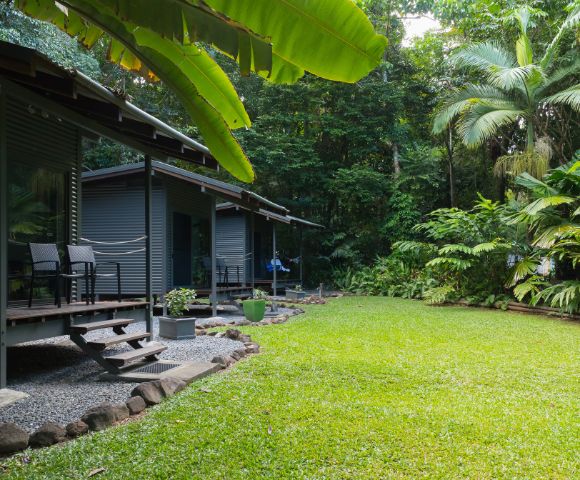 A serene garden area features two modern cabins with chairs on a wooden deck, surrounded by lush tropical vegetation and a well-maintained grassy lawn.