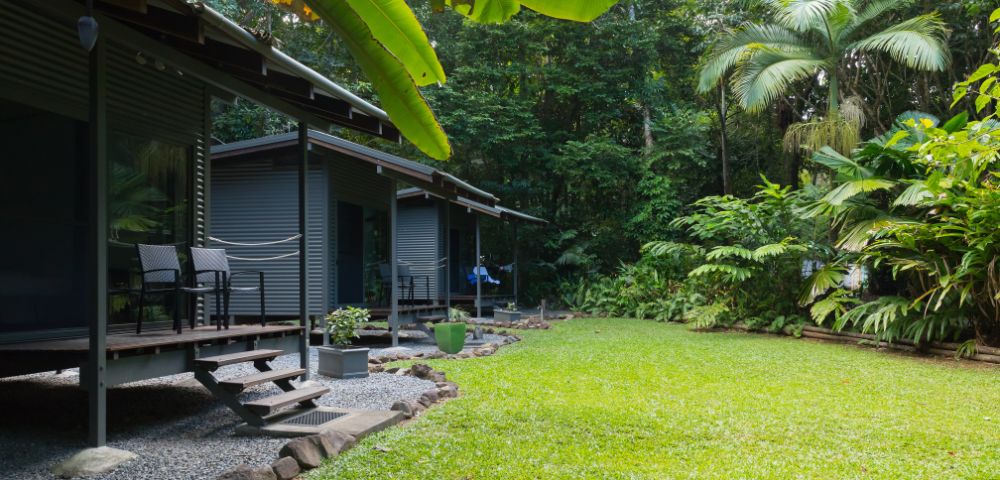 A serene garden area features two modern cabins with chairs on a wooden deck, surrounded by lush tropical vegetation and a well-maintained grassy lawn.