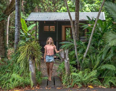 A woman stands on a wooden path surrounded by lush greenery, smiling as she approaches a rustic cabin nestled in a tropical forest.