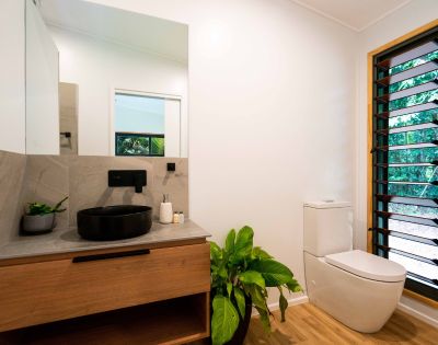 A modern bathroom featuring a dark circular sink on a grey countertop, wooden cabinetry, a white toilet, and a window with louvered shutters that shows lush greenery outside.