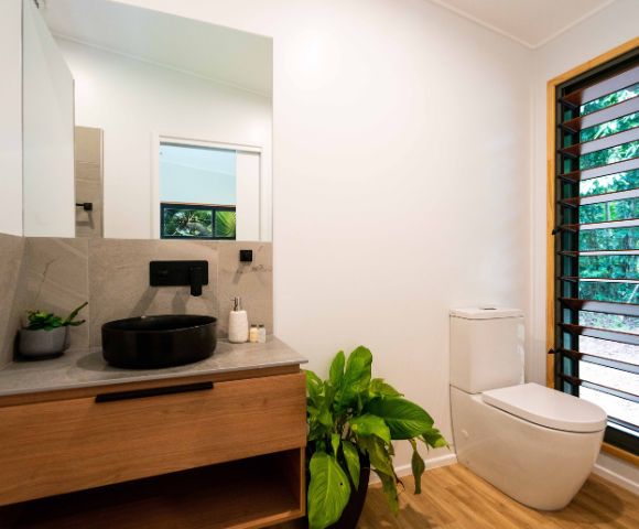 A modern bathroom featuring a dark circular sink on a grey countertop, wooden cabinetry, a white toilet, and a window with louvered shutters that shows lush greenery outside.