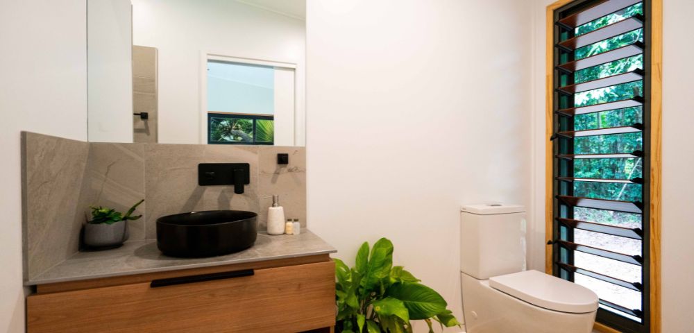 A modern bathroom featuring a dark circular sink on a grey countertop, wooden cabinetry, a white toilet, and a window with louvered shutters that shows lush greenery outside.