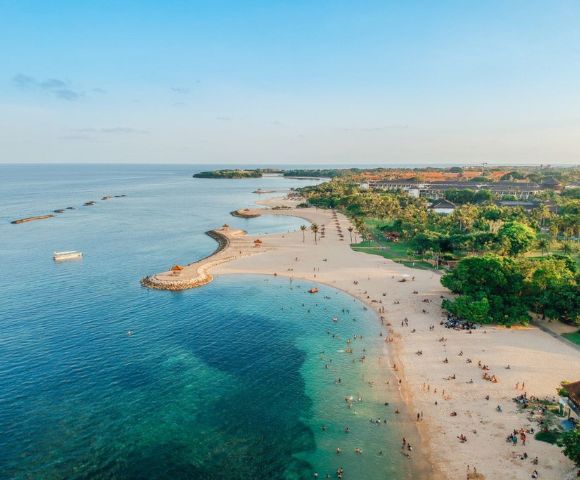 Aerial view of a serene beach with clear turquoise waters, scattered swimmers, sandy shores, and lush green trees. People relax under umbrellas, conveying a peaceful, sunny atmosphere.