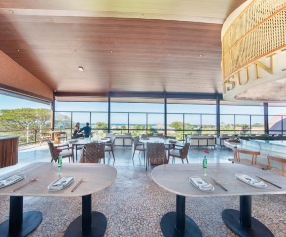 Bright restaurant interior with large windows showing a scenic view. Tables are set with chopsticks and bottles. Two people converse near a bar. Serene ambiance.