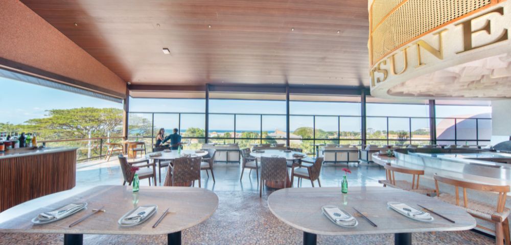 Bright restaurant interior with large windows showing a scenic view. Tables are set with chopsticks and bottles. Two people converse near a bar. Serene ambiance.