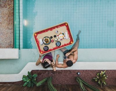 A couple in a pool share a floating breakfast tray adorned with flowers. Surrounded by plants, they enjoy a serene and romantic morning.
