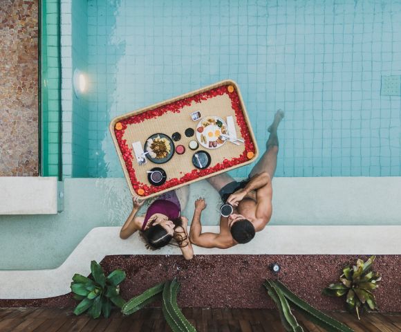 A couple in a pool share a floating breakfast tray adorned with flowers. Surrounded by plants, they enjoy a serene and romantic morning.