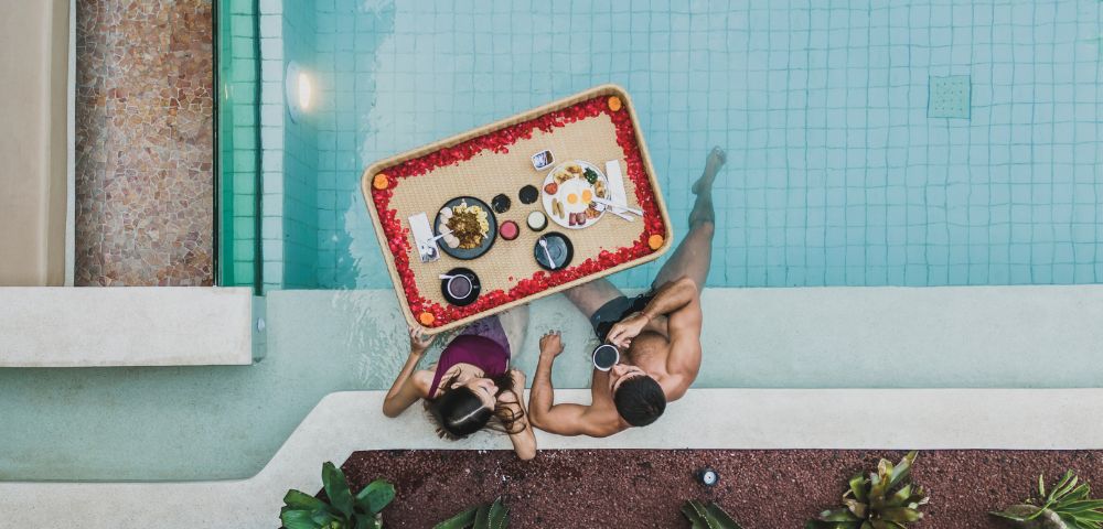 A couple in a pool share a floating breakfast tray adorned with flowers. Surrounded by plants, they enjoy a serene and romantic morning.