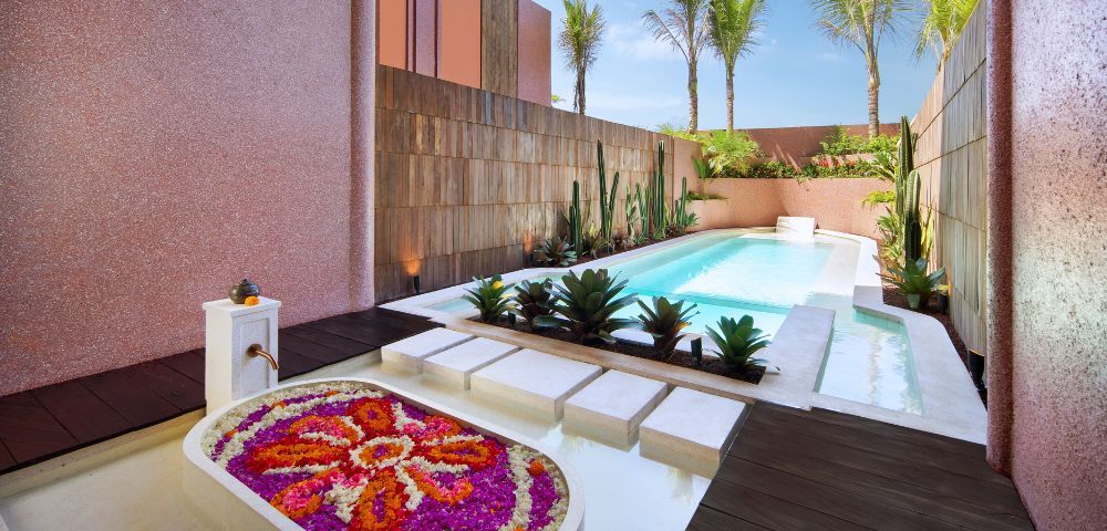 Luxurious outdoor patio featuring a sparkling pool surrounded by pink stucco walls, lush palm trees, and vibrant flowers, creating a serene oasis.