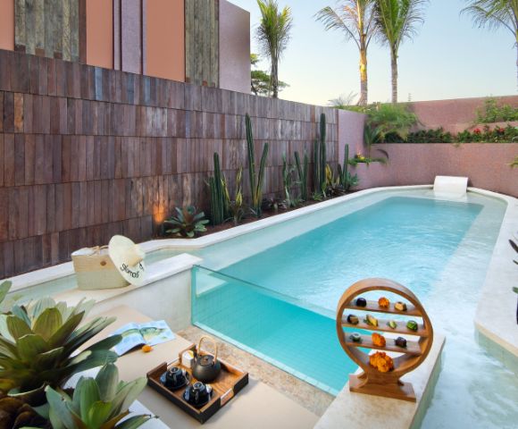A serene pool area with tropical plants and palm trees, featuring a small waterfall, wooden decor, and a stylish tea set, evoking a peaceful, luxurious vibe.