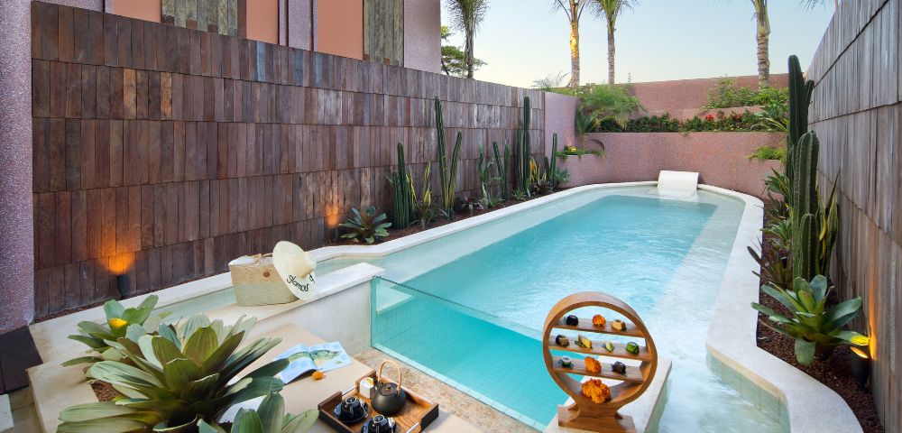 A serene pool area with tropical plants and palm trees, featuring a small waterfall, wooden decor, and a stylish tea set, evoking a peaceful, luxurious vibe.