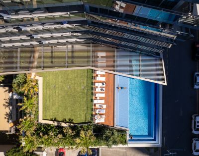 Aerial view of an urban rooftop featuring a lush green lawn, sun loungers, and a swimming pool. A modern glass building stands nearby, suggesting luxury and relaxation.