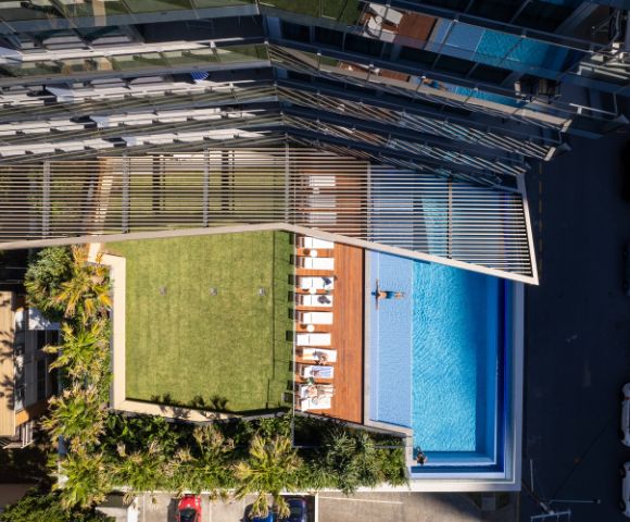 Aerial view of an urban rooftop featuring a lush green lawn, sun loungers, and a swimming pool. A modern glass building stands nearby, suggesting luxury and relaxation.