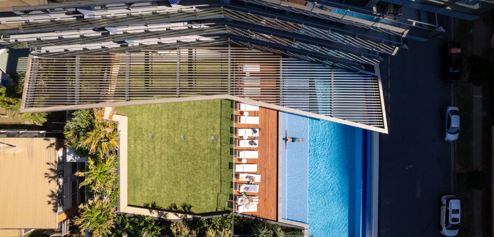 Aerial view of an urban rooftop featuring a lush green lawn, sun loungers, and a swimming pool. A modern glass building stands nearby, suggesting luxury and relaxation.