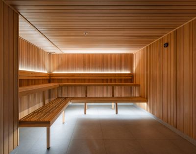 Modern sauna interior with warm wooden panels and bench seating. Subtle ambient lighting creates a calm, relaxing atmosphere. Tiled floor visible.