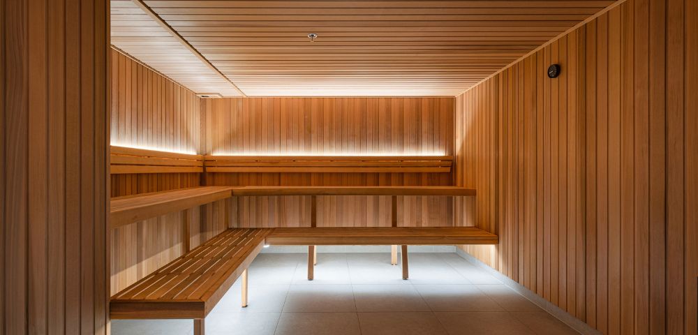 Modern sauna interior with warm wooden panels and bench seating. Subtle ambient lighting creates a calm, relaxing atmosphere. Tiled floor visible.