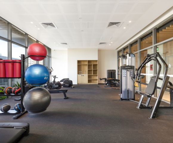 Modern gym with exercise equipment, including stability balls, weighted balls, and machines. Bright, spacious interior with large windows. Calm and inviting.