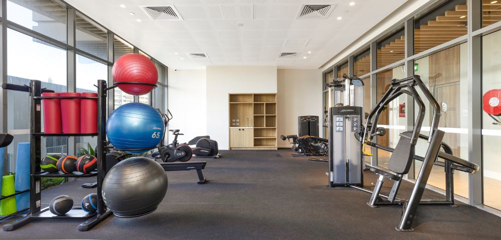 Modern gym with exercise equipment, including stability balls, weighted balls, and machines. Bright, spacious interior with large windows. Calm and inviting.