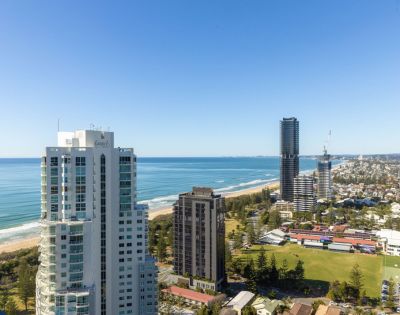 Aerial view of coastal cityscape with tall buildings along the ocean, lush greenery, and a clear blue sky. The scene conveys tranquility and urban vibrancy.