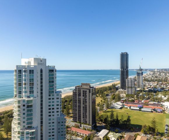 Aerial view of coastal cityscape with tall buildings along the ocean, lush greenery, and a clear blue sky. The scene conveys tranquility and urban vibrancy.