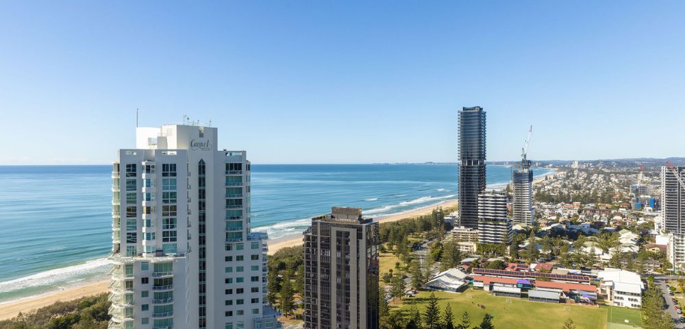 Aerial view of coastal cityscape with tall buildings along the ocean, lush greenery, and a clear blue sky. The scene conveys tranquility and urban vibrancy.