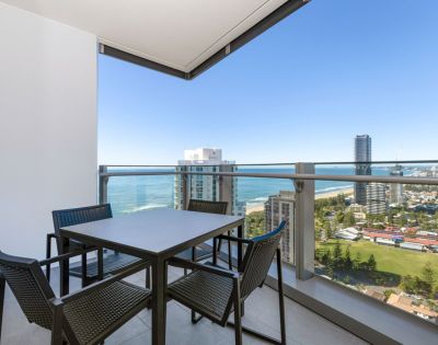 Balcony view featuring a modern outdoor dining table with four chairs. The backdrop includes city buildings, ocean, and a clear blue sky, evoking a serene ambiance.