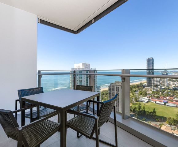 Balcony view featuring a modern outdoor dining table with four chairs. The backdrop includes city buildings, ocean, and a clear blue sky, evoking a serene ambiance.