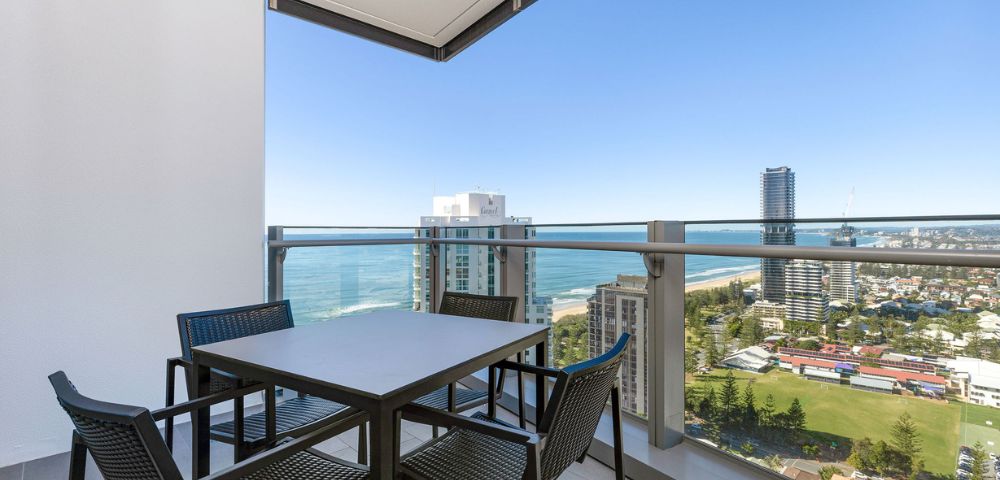 Balcony view featuring a modern outdoor dining table with four chairs. The backdrop includes city buildings, ocean, and a clear blue sky, evoking a serene ambiance.