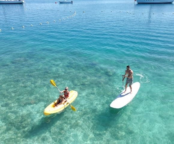 A woman paddles a yellow kayak while a man stands on a paddleboard, both enjoying the clear, turquoise water and sunny day.