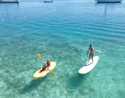A woman paddles a yellow kayak while a man stands on a paddleboard, both enjoying the clear, turquoise water and sunny day.