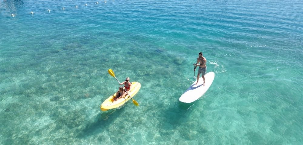 A woman paddles a yellow kayak while a man stands on a paddleboard, both enjoying the clear, turquoise water and sunny day.