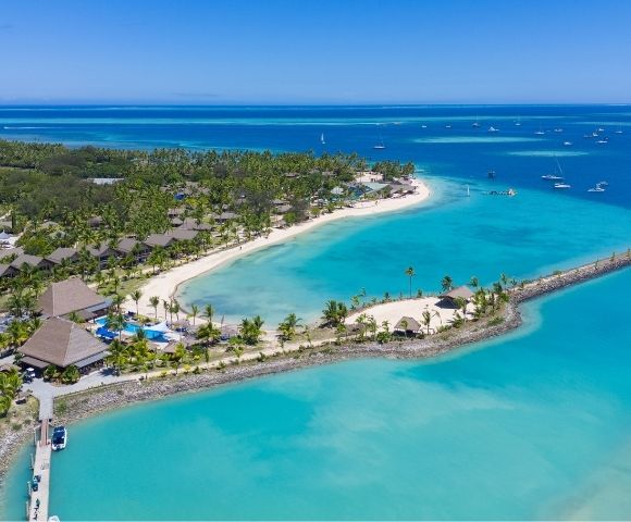 Aerial view of a tropical resort with lush greenery, sandy beaches, and turquoise waters, dotted with boats under a clear blue sky.
