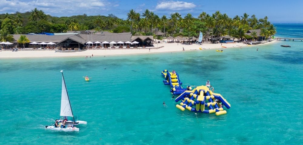 Aerial view of a tropical beach with clear turquoise water, inflatable water park, sailboat, and palm trees in the background.