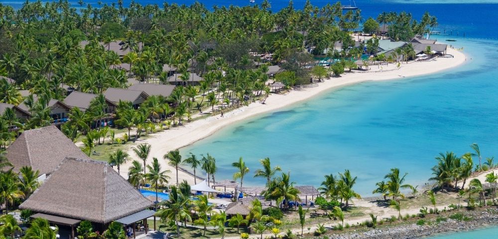 Aerial view of a tropical beach resort with palm trees, clear blue waters, and white sand, under a bright blue sky.