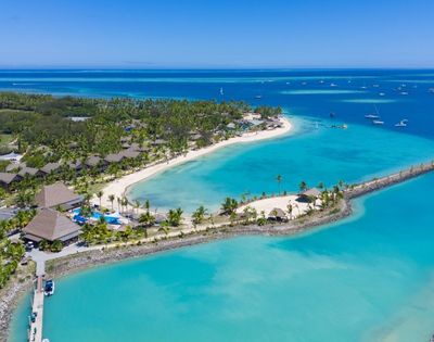 Aerial view of a tropical resort with lush greenery, sandy beaches, and turquoise waters, dotted with boats under a clear blue sky.