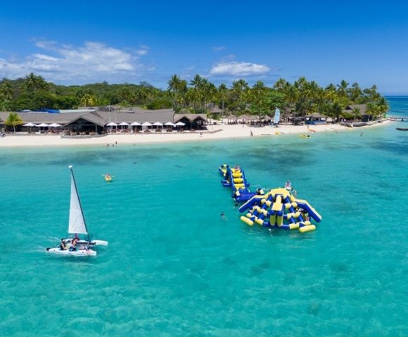 Aerial view of a tropical beach with clear turquoise water, inflatable water park, sailboat, and palm trees in the background.