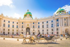 Horse-drawn carriage passes in front of a grand, white historic building with green domes under a bright blue sky. Tourists walk nearby. Elegant, lively scene.