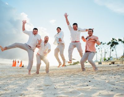 Five men in light-colored attire joyfully jump on a sandy beach at sunset. In the background, people in orange dresses, trees, and a cloudy sky are visible.